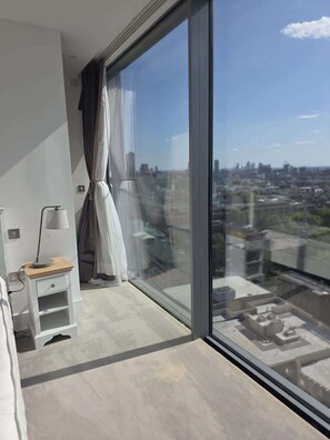 Ein modernes Schlafzimmer mit großem Fenster, Nachttisch mit Lampe und Blick auf eine Stadtansicht.