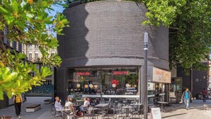 Un café al aire libre con mesas y sillas, un muro de ladrillo y un letrero para bar de cócteles y cafetería.