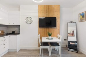A modern kitchen with white cabinets, a wooden accent wall, a flat-screen TV, a dining table with chairs, and a large mirror. - Amazing 1 bed - St John’s Wood (Greater London)