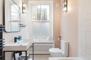 A modern bathroom with a white sink, toilet, and bathtub, a window with frosted glass, and wall-mounted light fixtures. - Stunning 2BR/2BA-ClaphamCommon (Greater London)