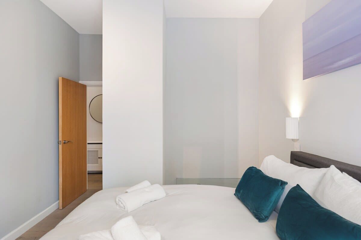 A hotel room with a bed, a nightstand, a wall-mounted lamp, and a door leading to a bathroom.