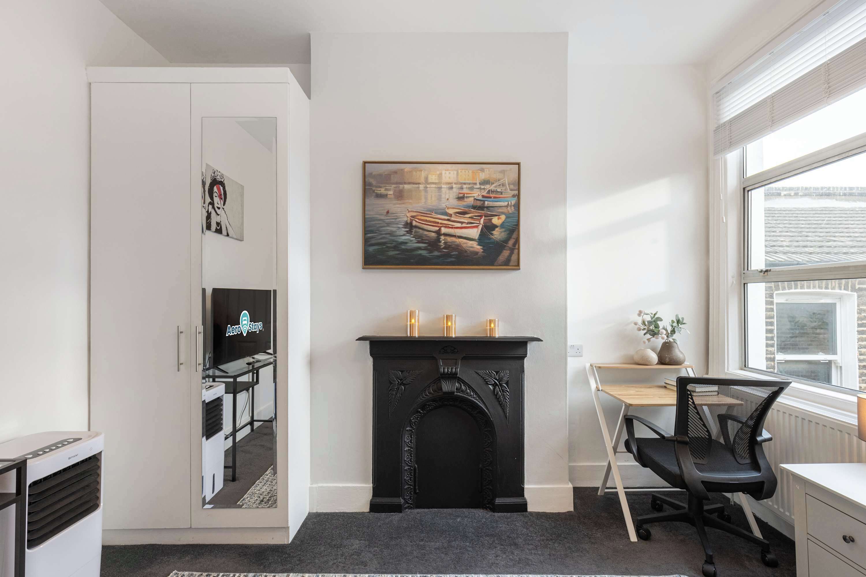 A room with a fireplace, a desk, a chair, a window, and a painting on the wall.