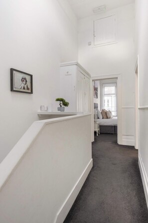 A narrow hallway with a staircase, a small seating area with a bed, and a framed picture on the wall. - Chic 2Bed w/Balcony - Kensal Rise (Greater London)