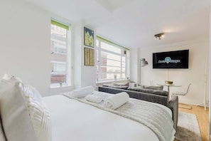 A modern hotel room with a bed, two seating areas, a flat-screen TV, and a window with blinds. - Superb 1BR w/AC - Chelsea (Greater London)