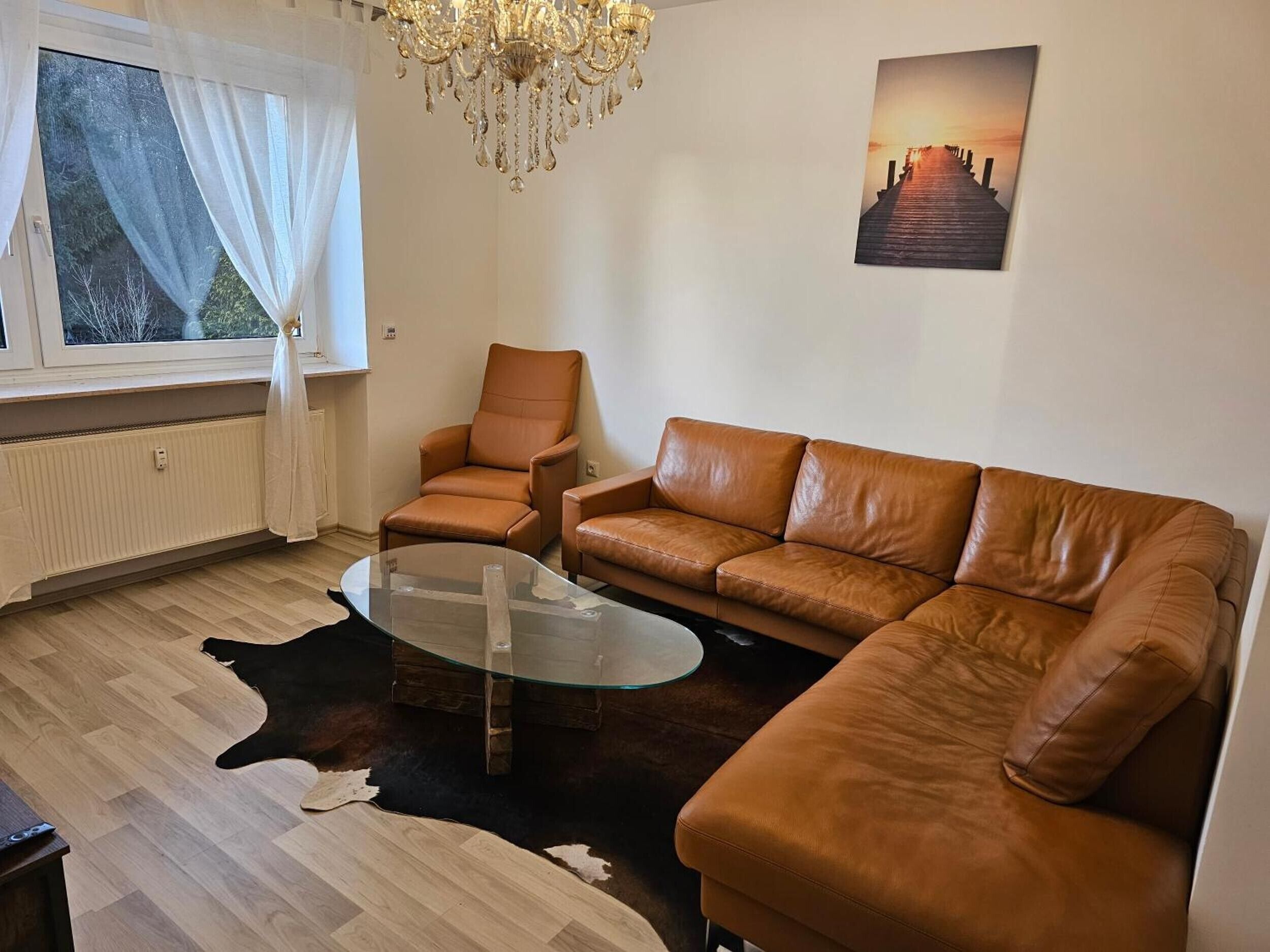 Ein Wohnzimmer mit einer Ledersofa, einem Glastisch, einer Chandelüster, einem Fenster mit transparenten Vorhängen und einem Gemälde an der Wand.