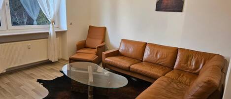 A living room with a leather sofa, a glass coffee table, a chandelier, a window with sheer curtains, and a painting on the wall.