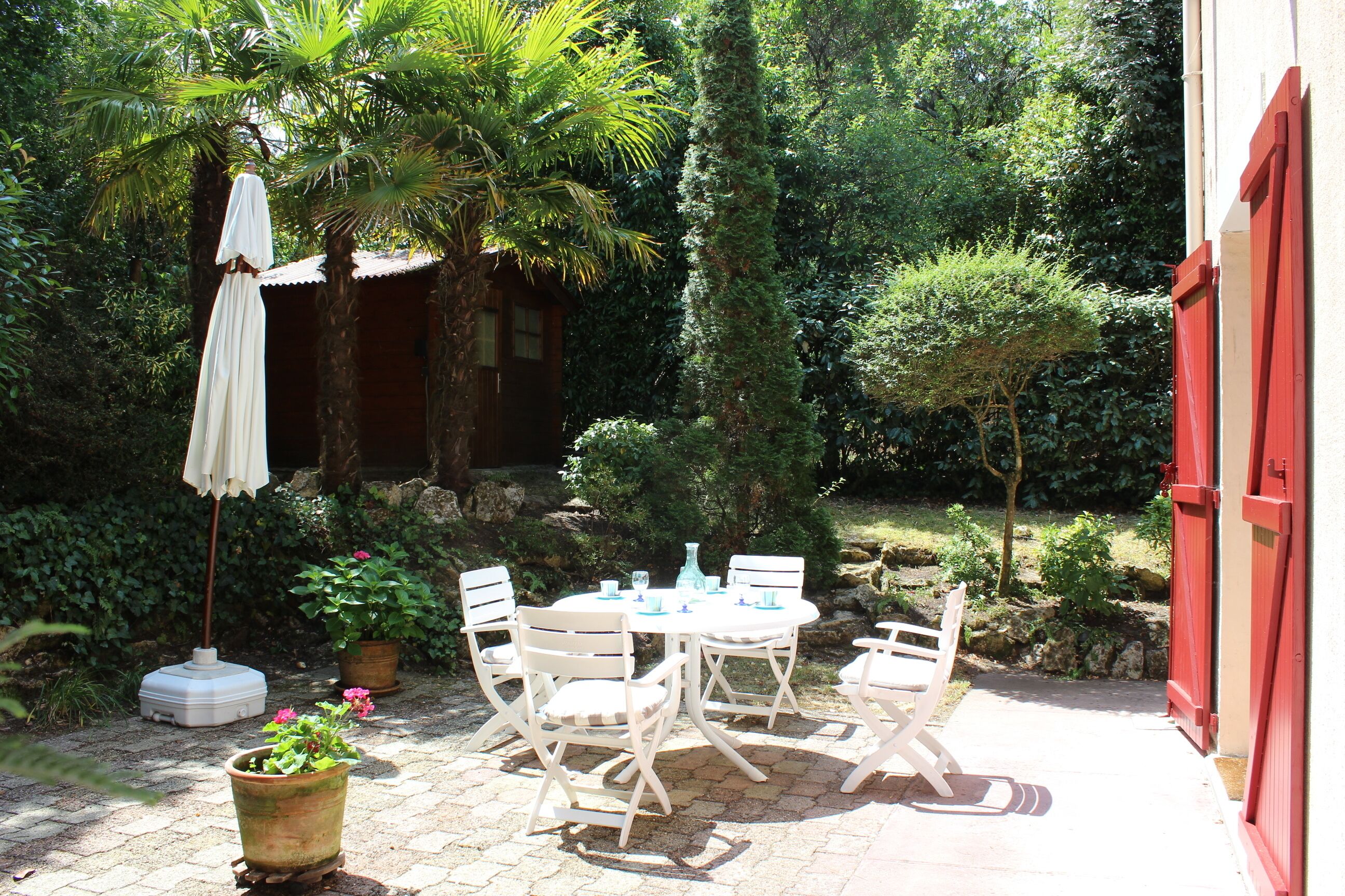 Une terrasse avec une table et des chaises, entourée de verdure et bordée d’un bâtiment rouge.