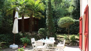 Eine Terrasse mit Tisch und Stühlen, umgeben von Pflanzen und einem roten Gebäude.