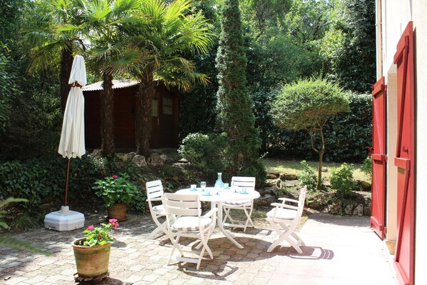 Eine Terrasse mit Tisch und Stühlen, umgeben von Pflanzen und einem roten Gebäude.