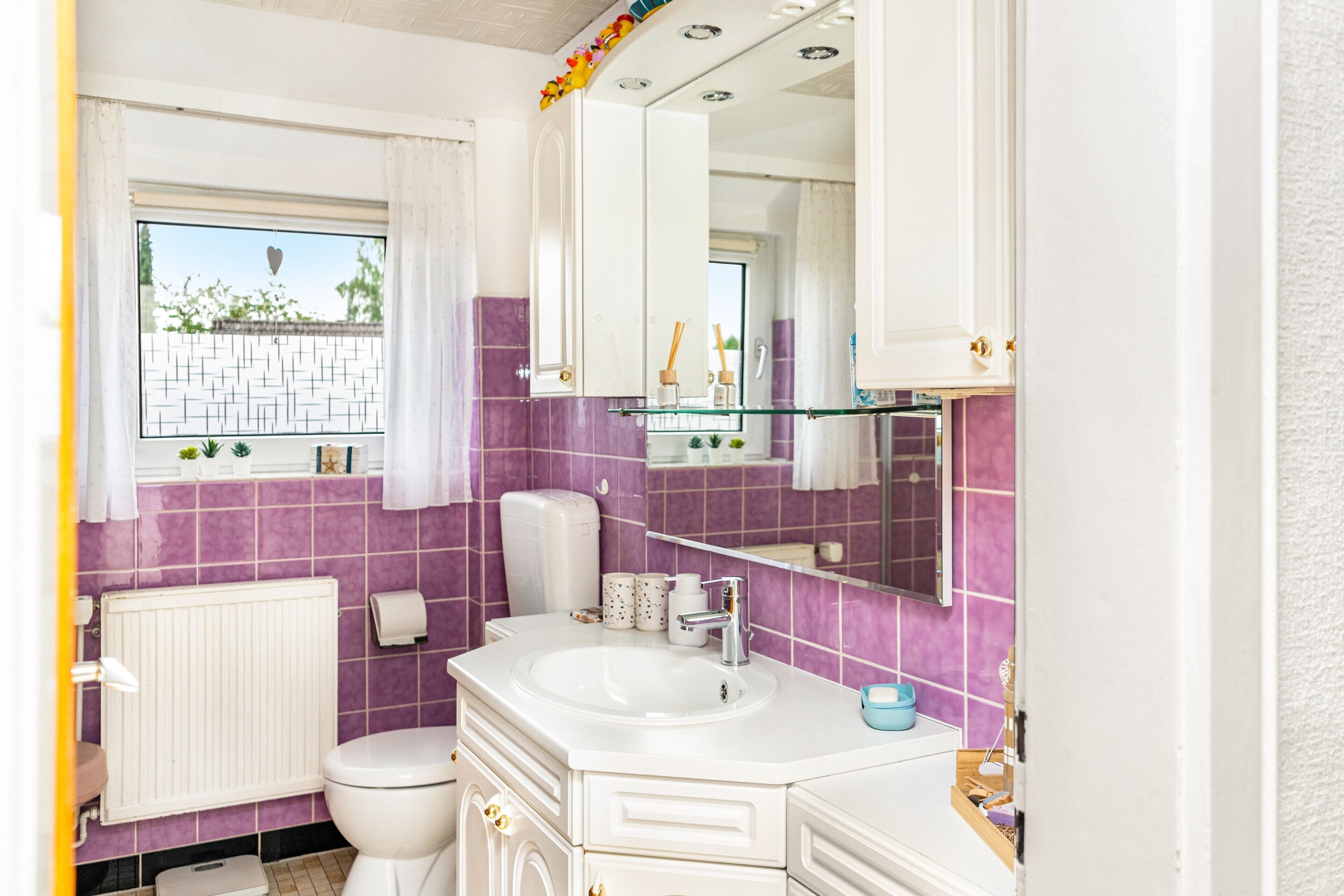 A bathroom with a white sink, a toilet, a mirror, and a window with a view of greenery outside.