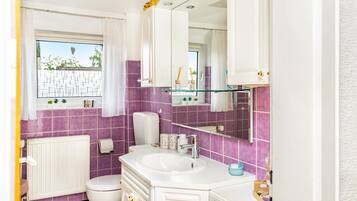 A bathroom with a white sink, a toilet, a mirror, and a window with a view of greenery outside.