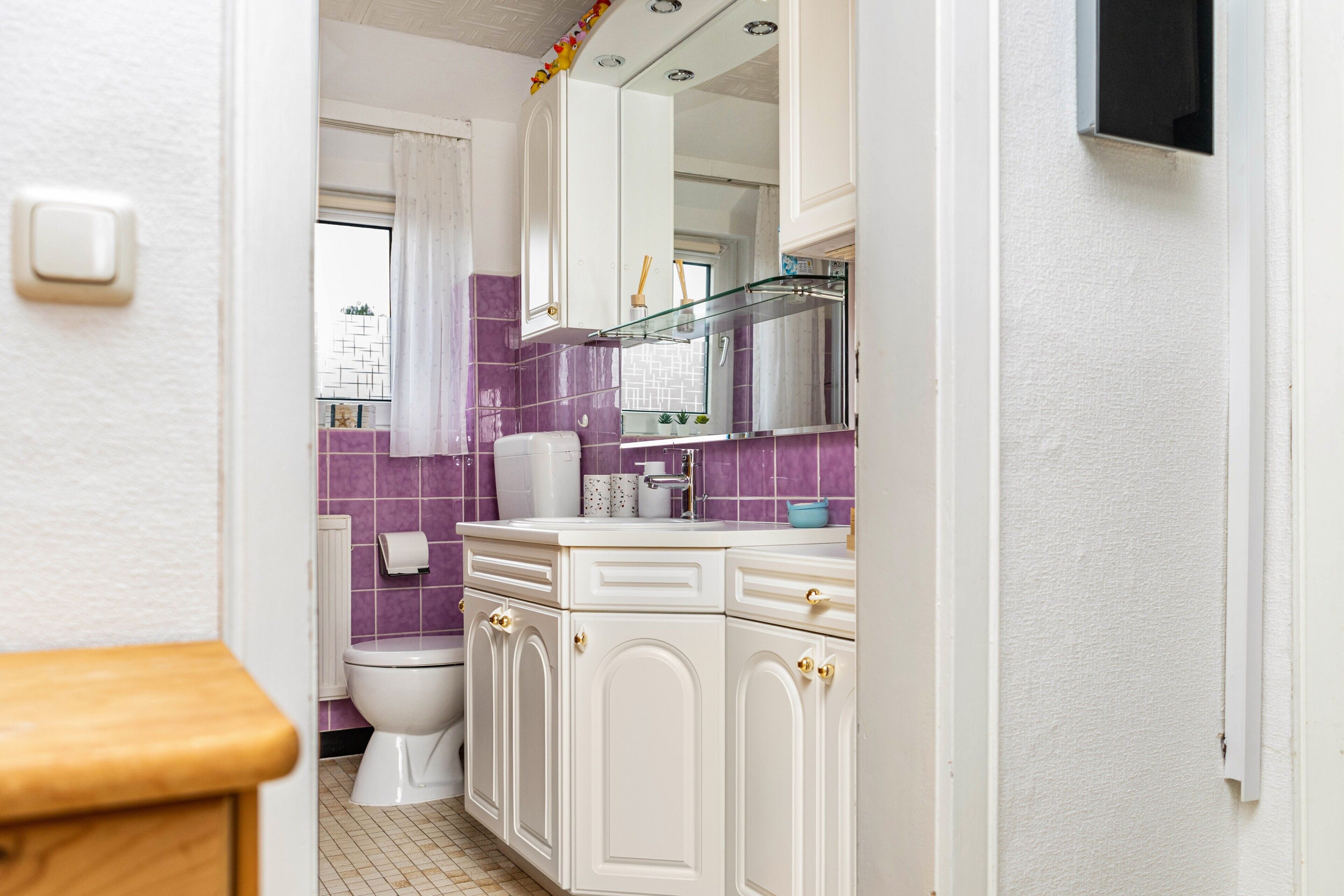 Een badkamer met toilet, wastafel, spiegel en paars betegelde wanden.
