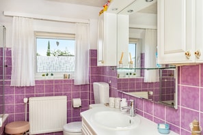 Ein Badezimmer mit einem weißen Waschbecken, einer Toilette, einem Fenster mit weißen Vorhängen und violetten Fliesen.