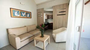 Een woonkamer met een beige bank, een klein houten bijzettafeltje met een vaas, en een witte fauteuil. Op de achtergrond is een keuken zichtbaar.