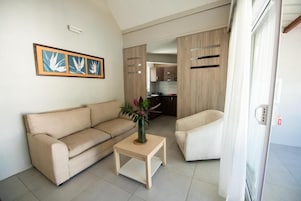 Una sala de estar con un sofá beige, una pequeña mesa de centro de madera con un jarrón y un sillón blanco. Se aprecia una cocina al fondo.