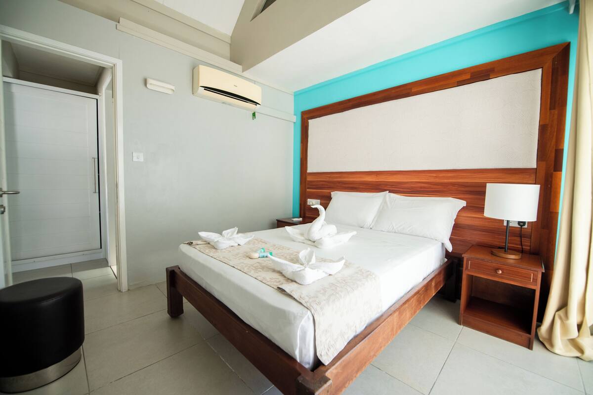Une chambre d’hôtel avec un lit en bois, une table de chevet avec une lampe, un appareil de climatisation et une porte.
