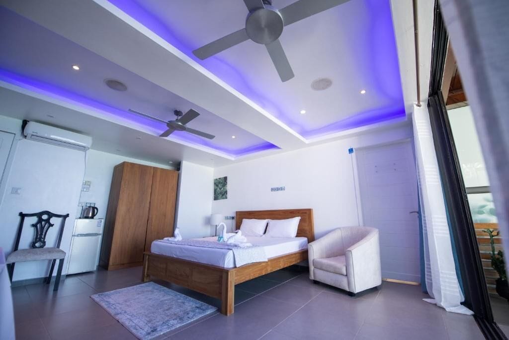 Un dormitorio moderno con cama, una silla, ventilador de techo y una pequeña kitchenette.