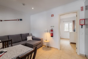 A modern living room with a grey sofa, a dining table with patterned tablecloth, a standing lamp, and a fire extinguisher on the wall. - Apartment 'Cottage 1' with Shared Pool, Wi-Fi and Air Conditioning (Alcácer do Sal)