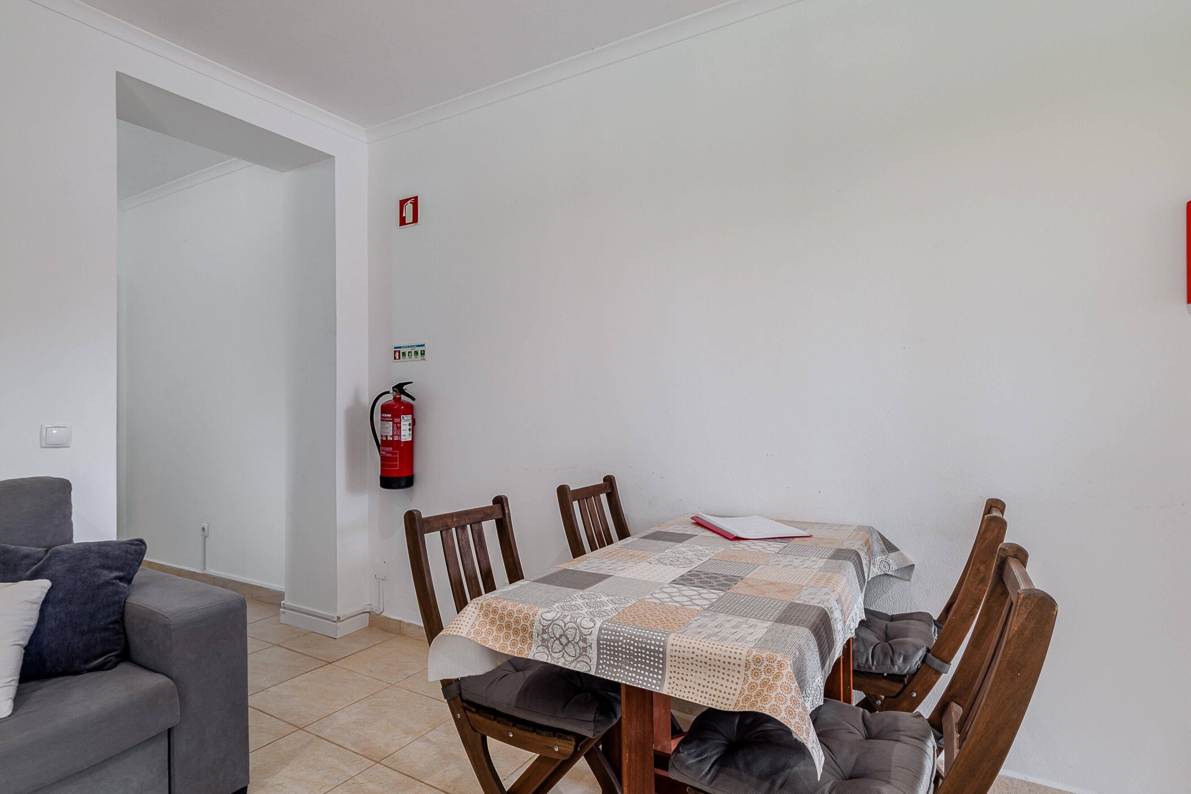 Een eetgedeelte met een tafel bedekt met een tafelkleed met een patroon, houten stoelen, een brandblusser aan de muur en een grijze bank.