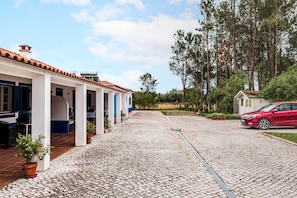 Une cour avec un chemin pavé de pierres, une voiture rouge et un bâtiment doté d’un toit en tuiles et de colonnes.