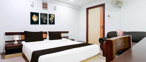A hotel room with a bed, a desk, a chair, a fan, and artwork on the wall.