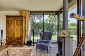 A room with a wooden wardrobe, a grey armchair, a fireplace, and a view of greenery outside. - Villa Greta with Mountain Views, Private Pool, and Wi-Fi (Vilanant)
