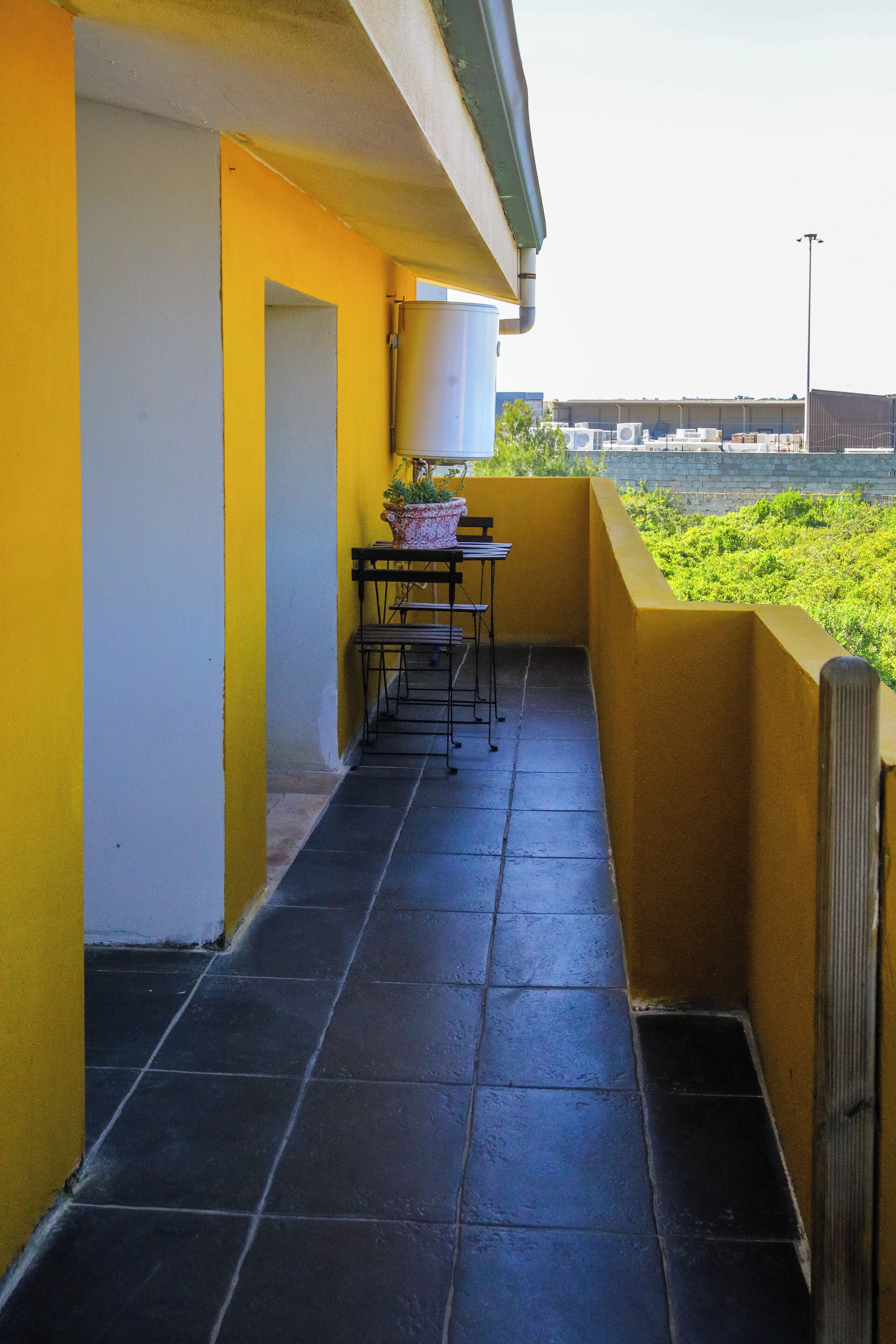 A narrow balcony with a tiled floor, a small table with chairs, and a potted plant.