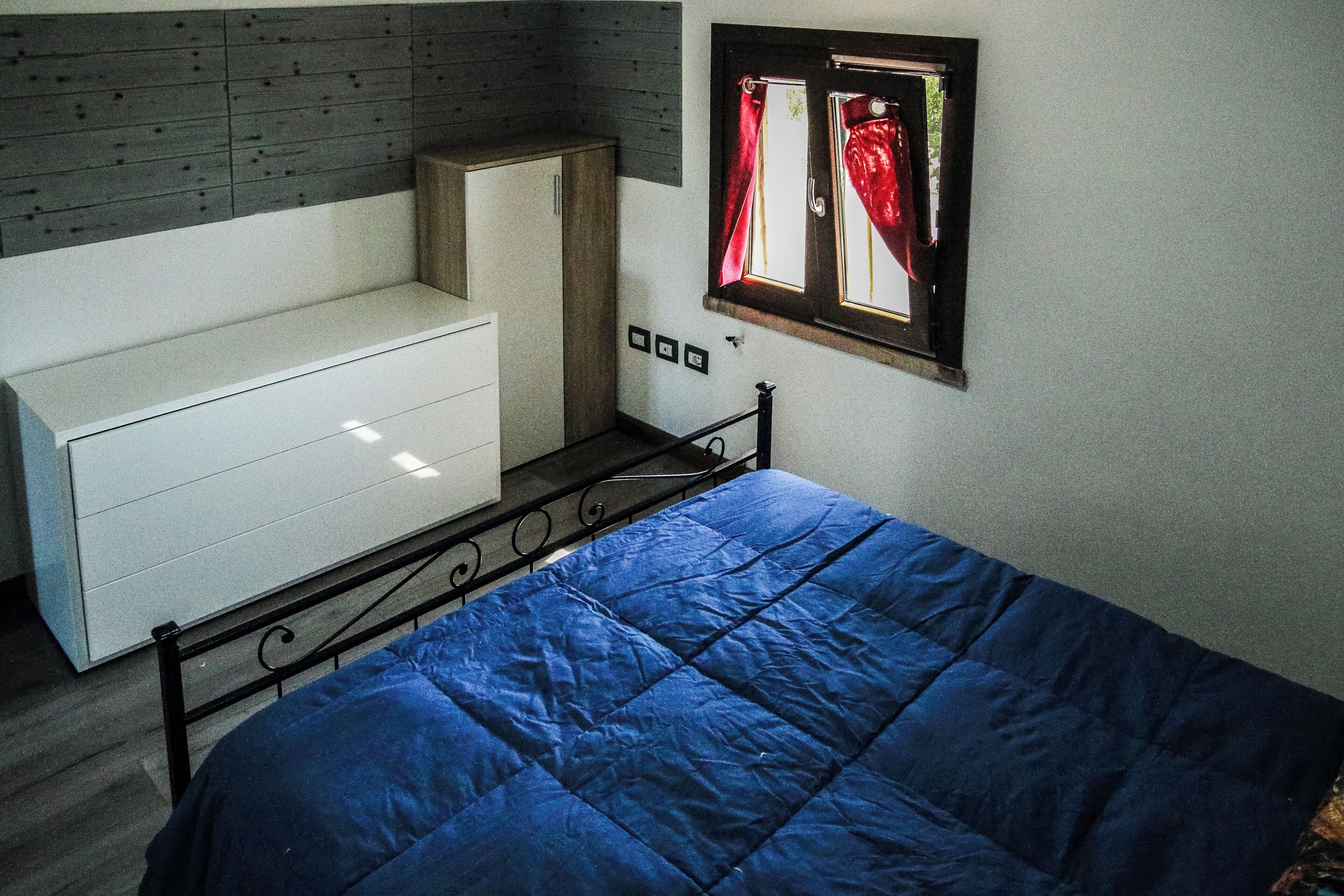 A bedroom with a bed, a white dresser, a window with curtains, and a wooden headboard.