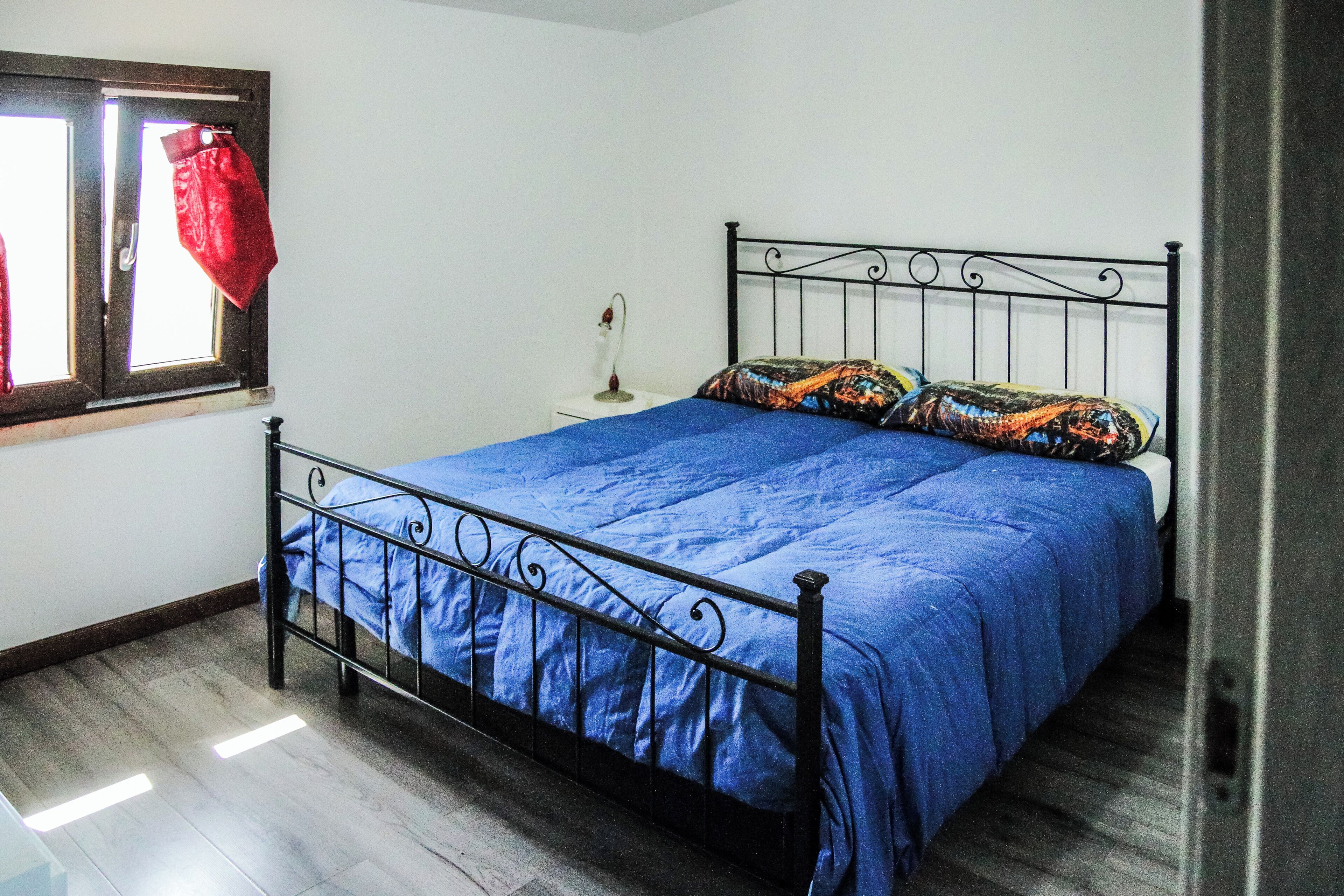 Un dormitorio con estructura de cama metálica, colcha azul, mesita de noche con lámpara y ventana con cortina roja.