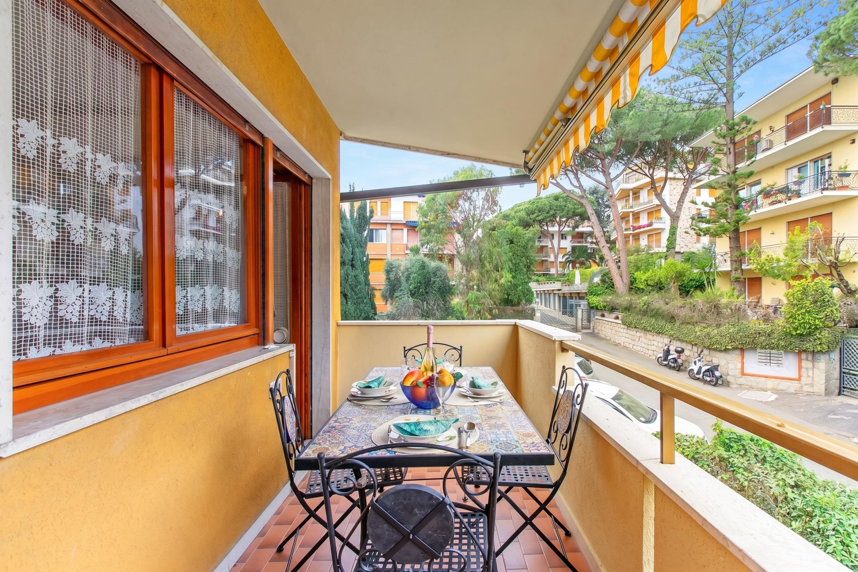 Varanda com mesa posta para refeições, uma cadeira e vista para a rua e edifícios.