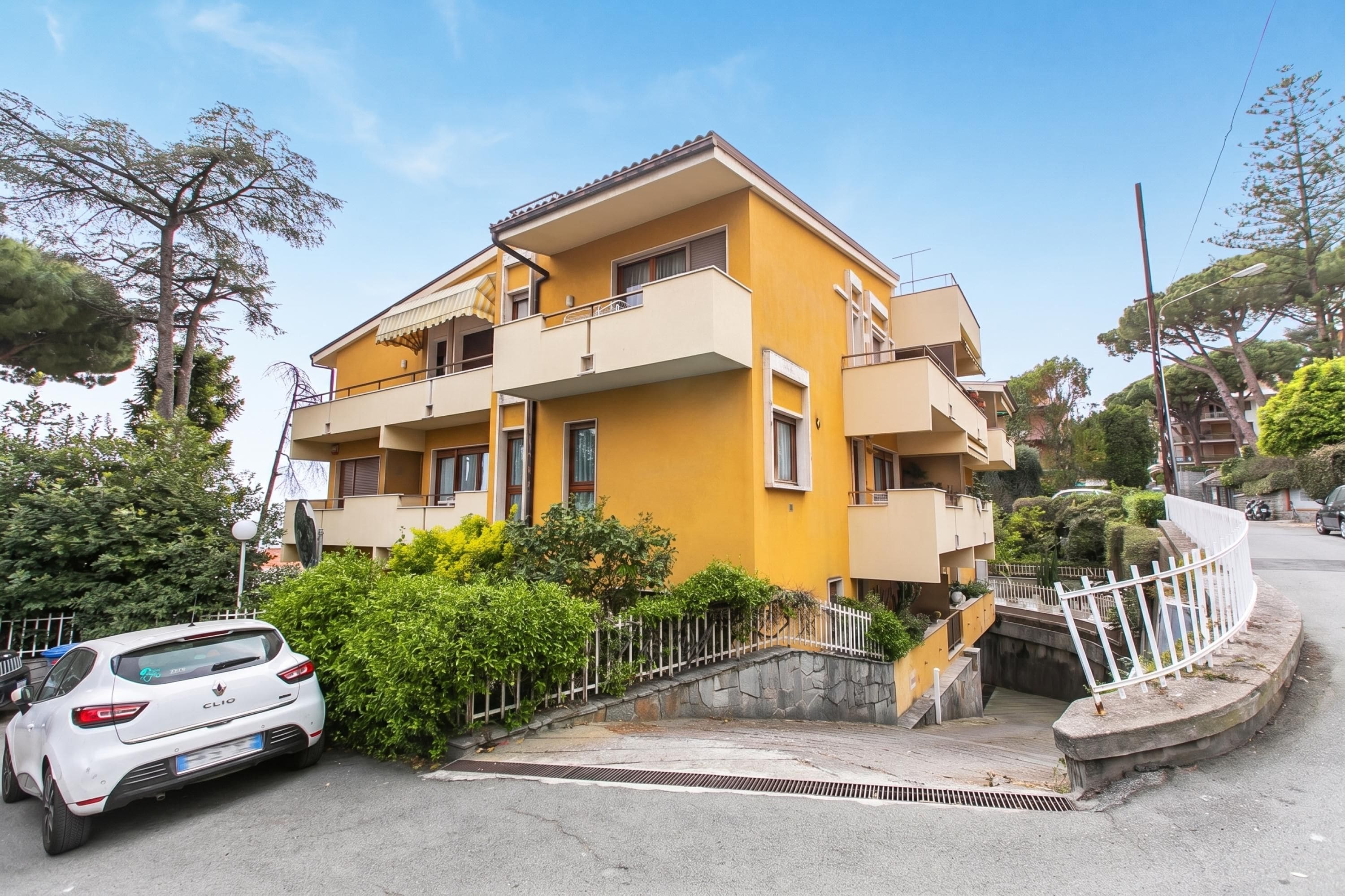 Um edifício residencial amarelo de vários andares com varandas, um carro branco estacionado em frente e uma cerca branca ao redor da propriedade.