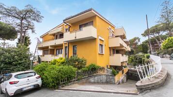Um edifício residencial amarelo de vários andares com varandas, um carro branco estacionado em frente e uma cerca branca ao redor da propriedade.