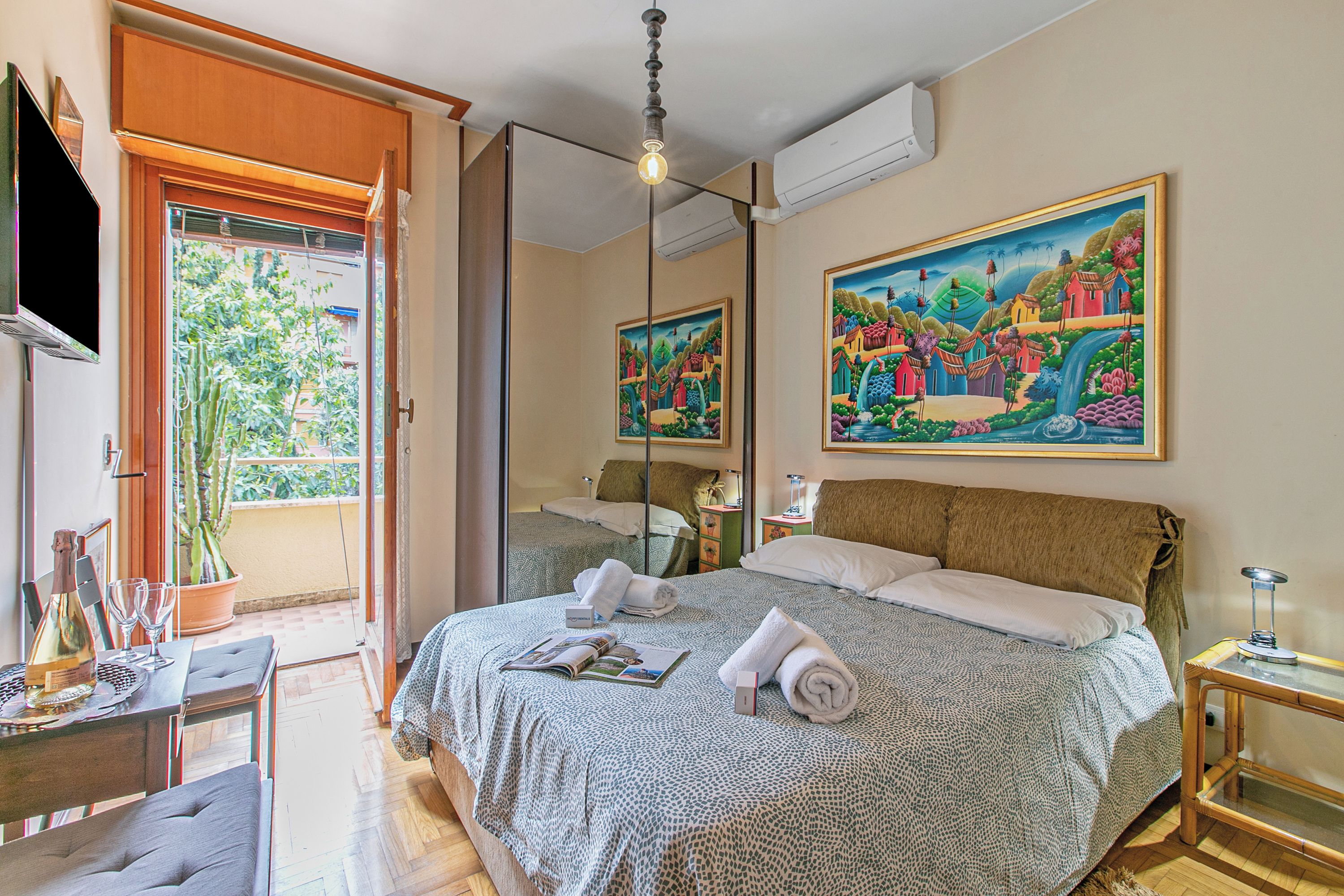 Quarto de hotel com uma cama, uma mesa com garrafa e copos, um espelho e um quadro na parede.