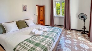 Un lit bien fait, recouvert d’une couverture à carreaux, un ventilateur sur pied, une fenêtre avec des rideaux et une porte donnant sur une autre pièce.