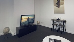 A modern living room with a flat-screen TV, a dining table with chairs, a small side table with a lamp, and a large abstract painting on the wall. - Apartment Sleeps 2 - Free Street Parking (Gateshead)