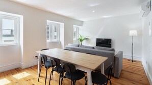 A modern living room with a wooden dining table, black chairs, a grey sofa, a flat-screen TV, and a potted plant. - Graça Duplex With A View by Homing (Lisbon)
