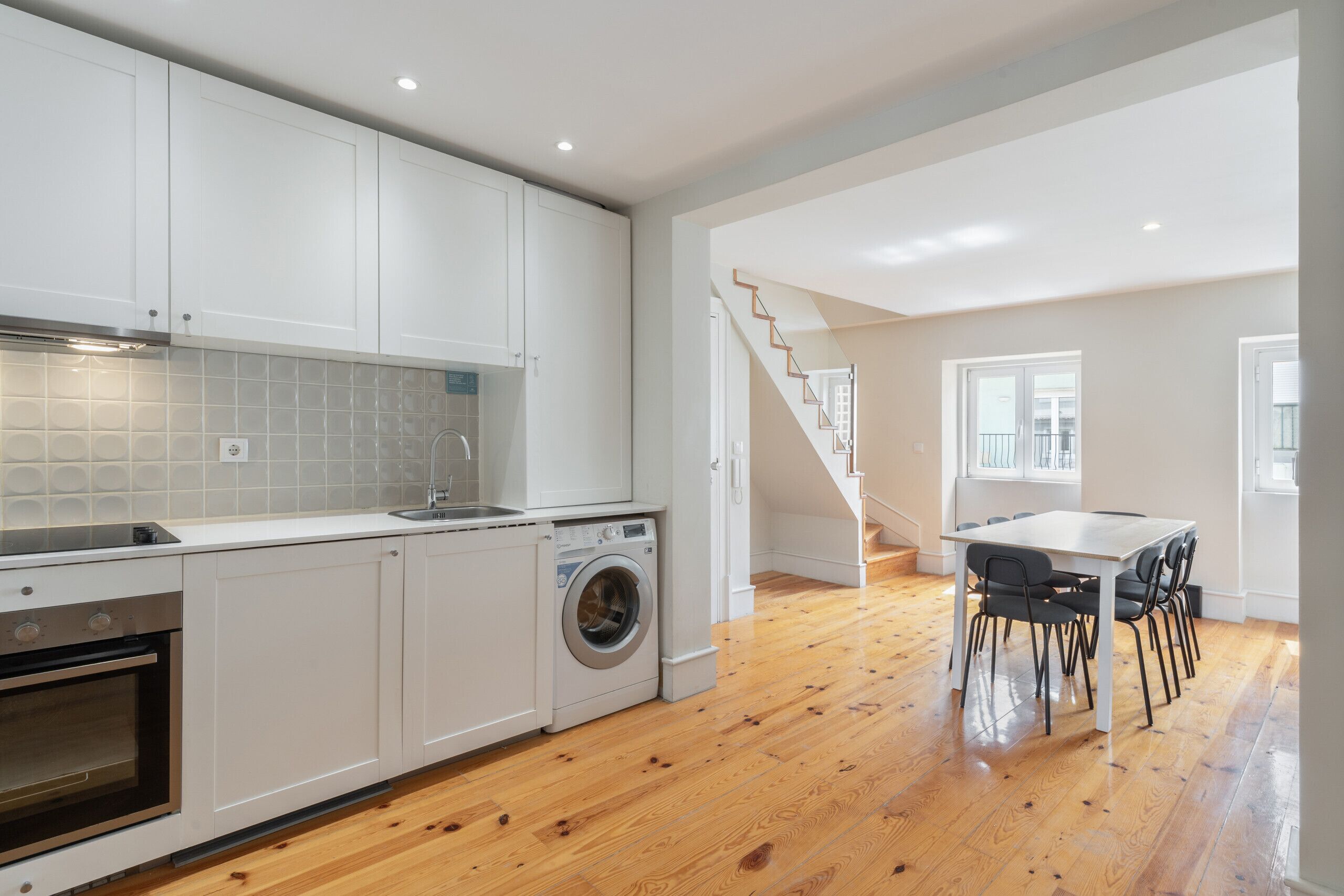 A modern kitchen with white cabinets, a built-in oven, a washing machine, and a dining area with a table and chairs.