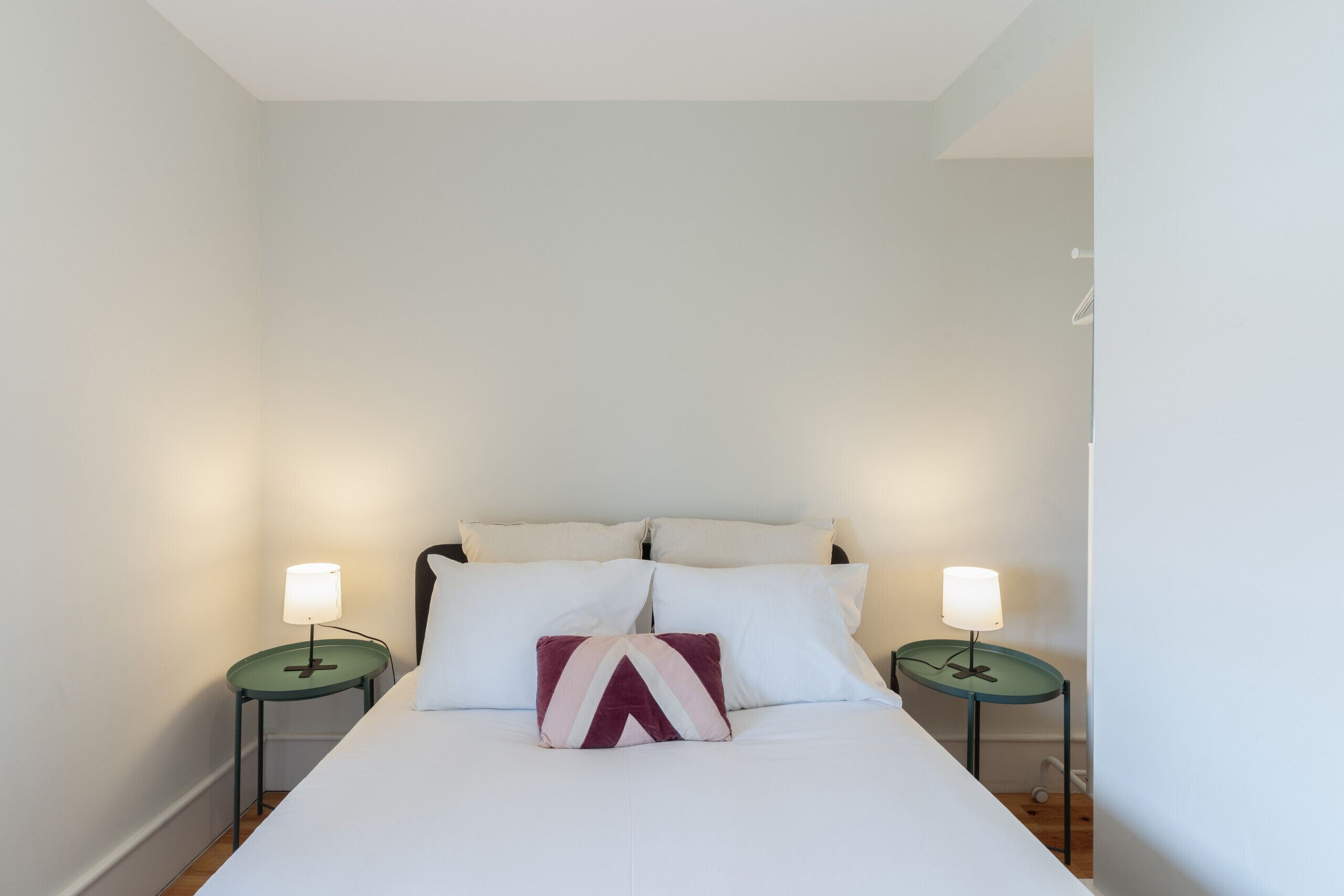 A neatly made bed with white bedding and a single decorative pillow, flanked by two bedside tables with lamps, in a simple room with white walls.