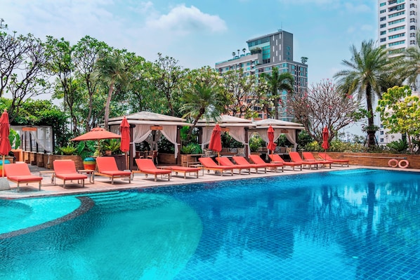A swimming pool with lounge chairs and umbrellas, surrounded by greenery and modern buildings.