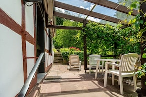 A covered patio with stone flooring, wooden beams, and white chairs.