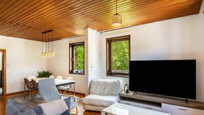 Una sala moderna con techo de madera, un televisor de pantalla plana grande, un sofá, un comedor con mesa y sillas, y un ventanal con vistas a zonas verdes.