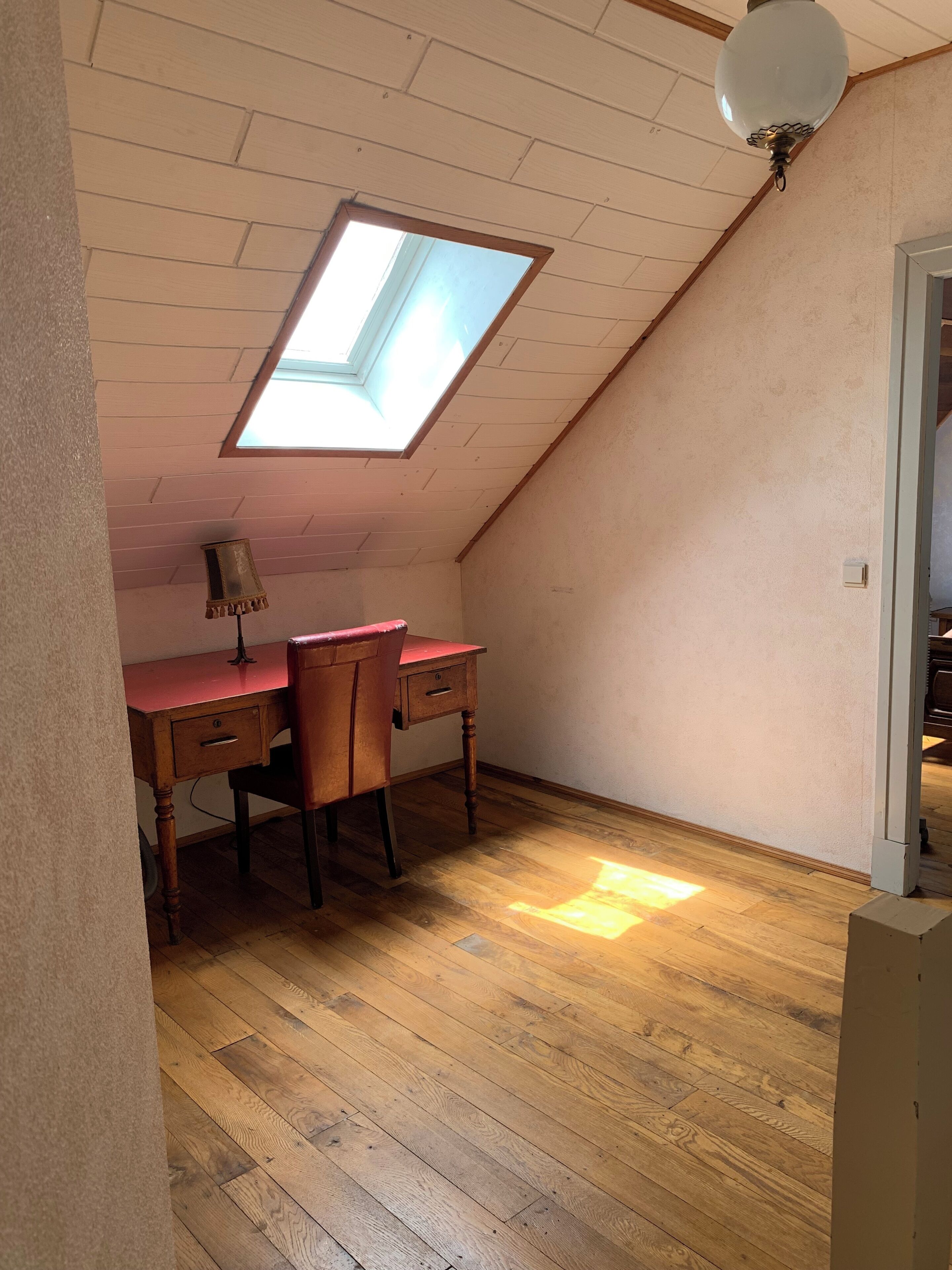 A room with a sloped ceiling, a skylight, a desk with a lamp, and wooden flooring.