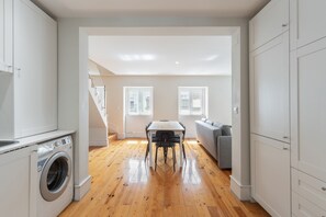 A modern kitchen with a built-in washer, a dining area with a table and chairs, and a gray sofa in a bright room with wooden flooring and white walls. - GRA A Duplex With A View by Homing (Lisbon)