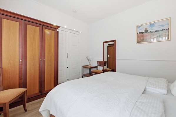 A bedroom with a large bed, wooden wardrobe, a small table with a chair, and a painting on the wall. - Villa Ascania, Room 1 in Zinnowitz - Historic flair meets vacation tranquillity (Zinnowitz)