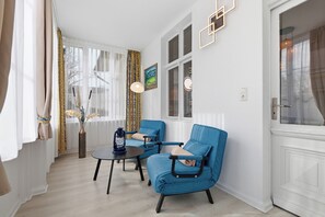 Ein Zimmer mit zwei blauen Sesseln, einem kleinen runden Tisch, einer Blumenvase und einem Gemälde an der Wand.