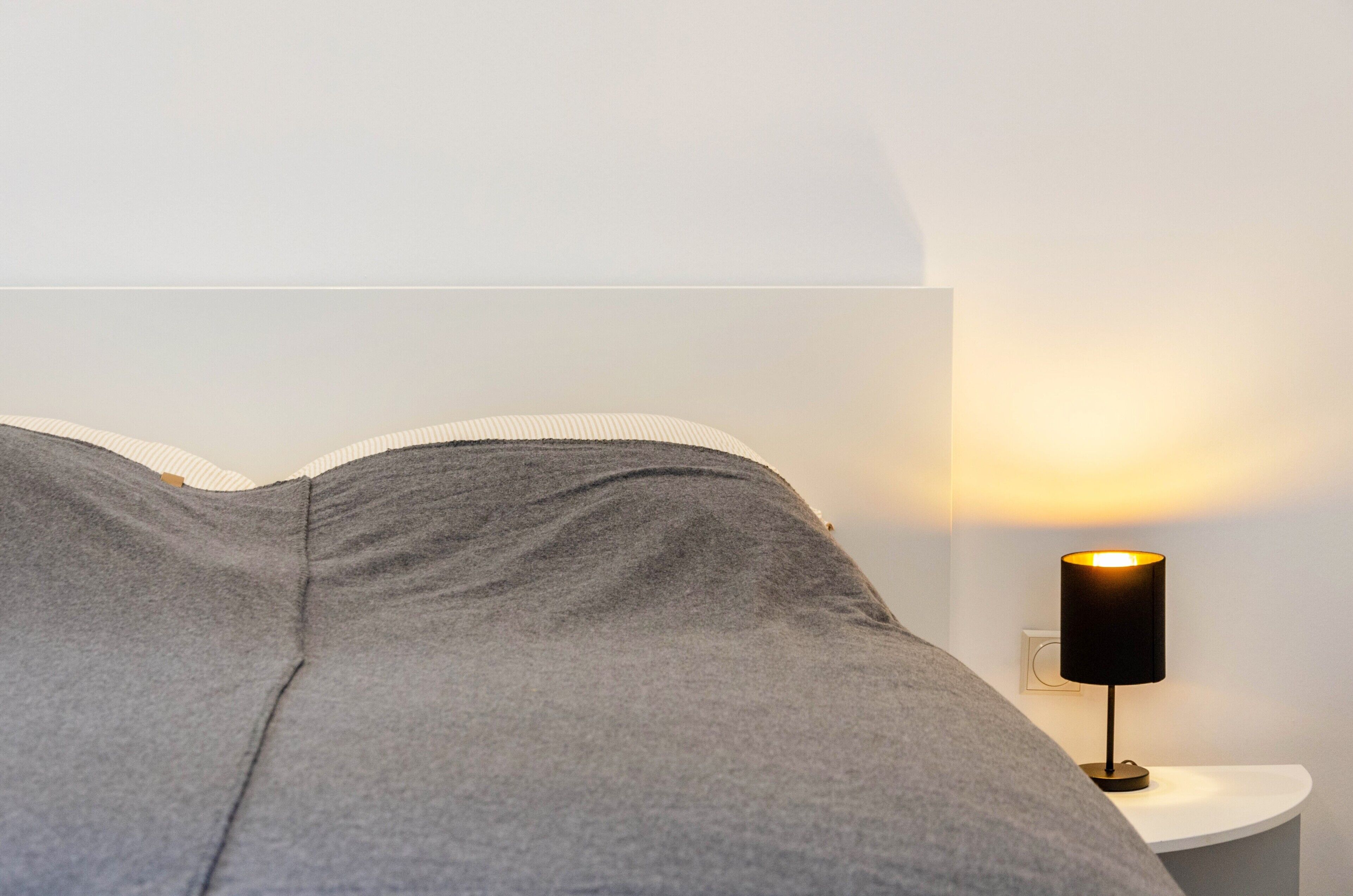 A bed with grey bedding, a white wall, and a nightstand with a black lamp.
