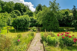 Ein gepflasterter Weg schlängelt sich durch einen üppigen Garten mit einer Bank und einem Tor.
