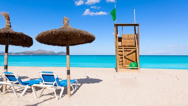 A beach with two thatched umbrellas, a lifeguard tower, and two blue lounge chairs. - Apartment Son Baulo Llimona (Ca´n Picafort)
