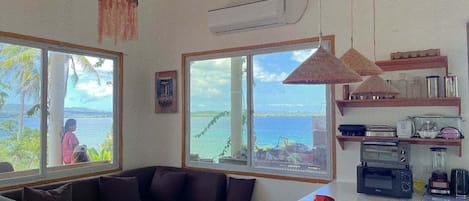 A room with a sofa, a dining table, and a view of the sea. There are pendant lights, a shelf with kitchen items, and an air conditioning unit.