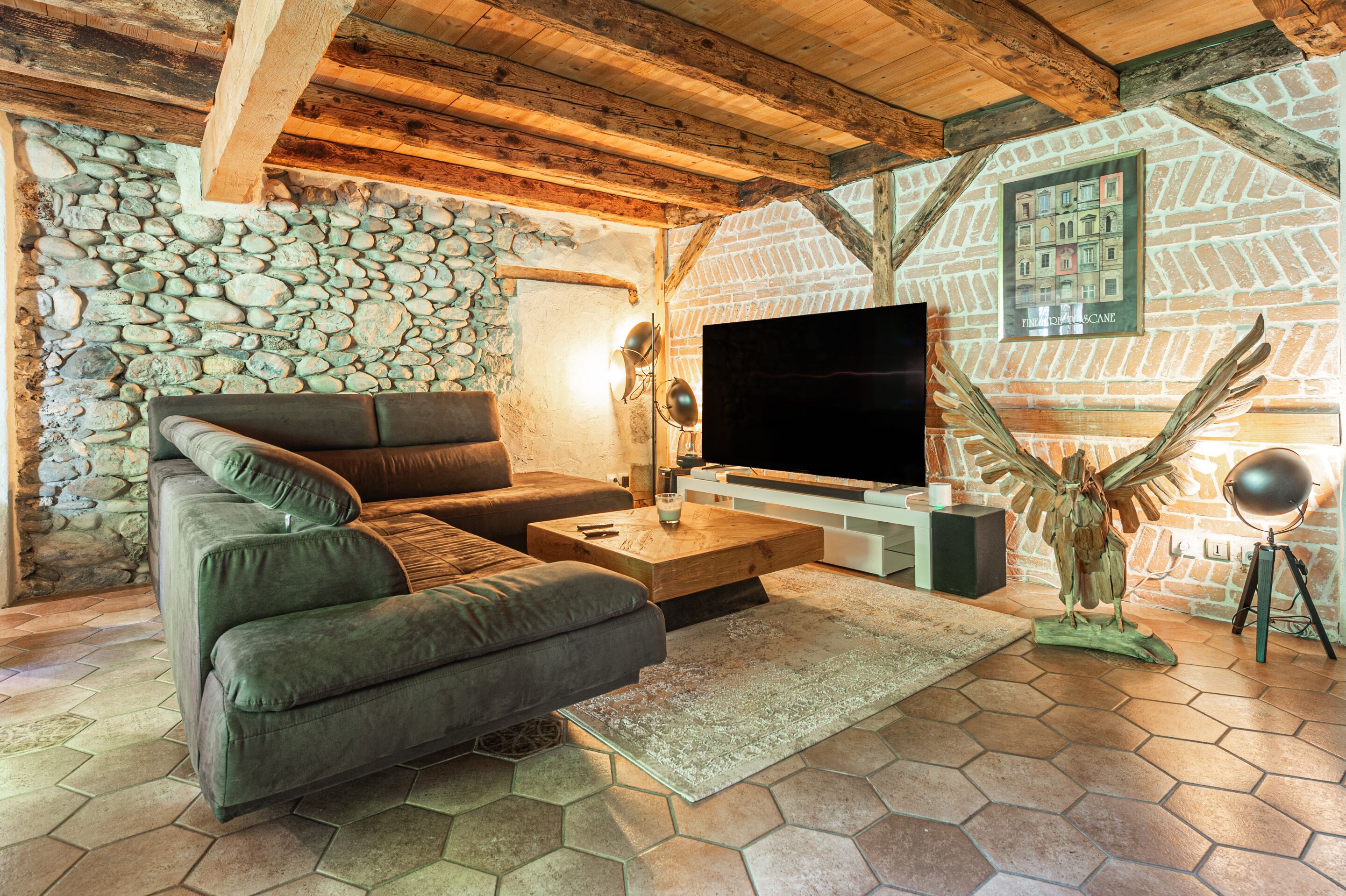 A cozy living room with a stone wall, wooden beams, a large flat-screen TV, a green sofa, and a decorative bird sculpture.